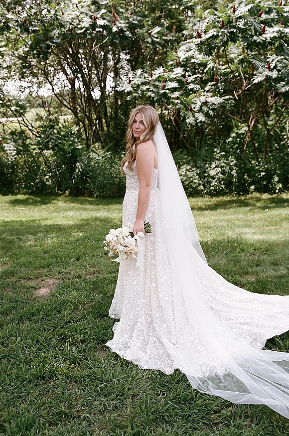 Bridal portrait of a bride holding bouquet in a strapless lace wedding dress with cathedral veil flowing across a green garden lawn