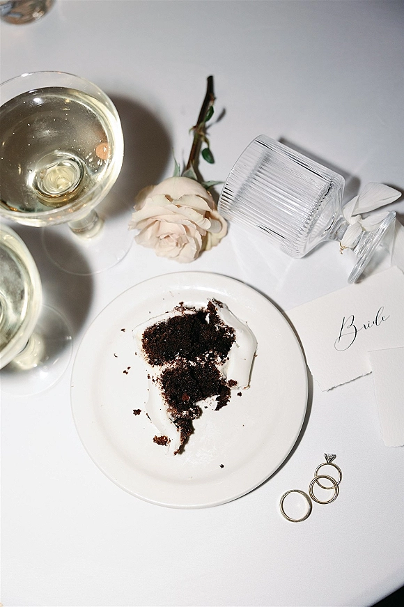 Wedding flat lay with wedding rings flat lay, champagne glasses, calligraphy place card, white rose, and cake slice on white tablecloth