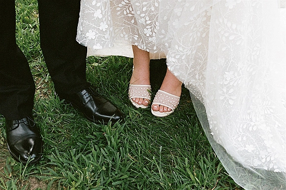 Wedding shoes beside groom black dress shoes on green grass lawn, bride in pearl bridal sandals with lace dress hem and tulle overlay
