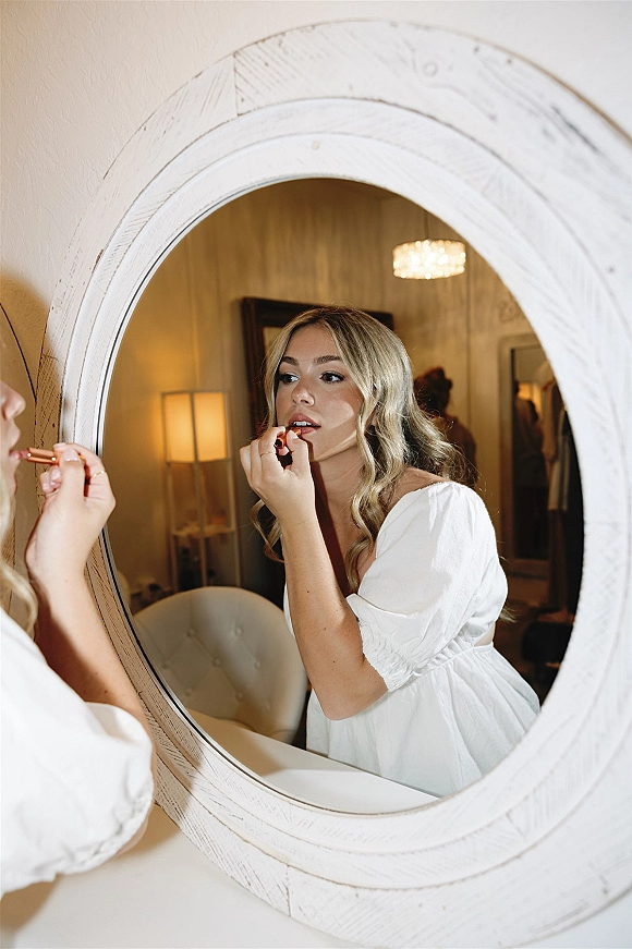 Bridal getting ready as bride applying lipstick in a white robe at the vanity, reflected in a round mirror under a chandelier in bedroom