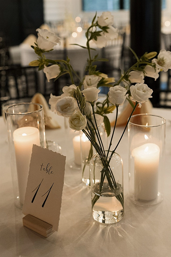 Reception table decor with bud vase centerpiece of white flowers, hurricane candle holders, and a calligraphy table number in window light