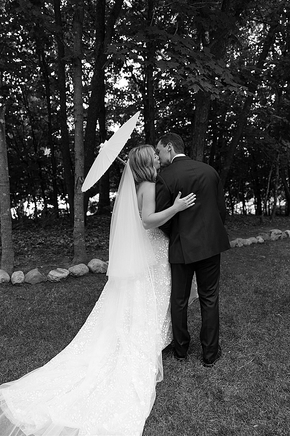 Wedding kiss portrait in black and white, bride and groom kissing from behind with long veil train, on a tree-lined lawn holding a white parasol