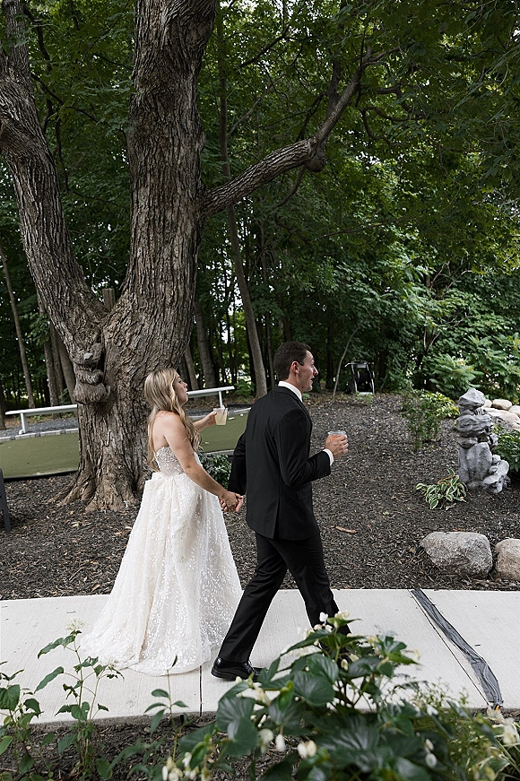 Wedding recessional as bride and groom walking away holding hands, strapless lace gown and tuxedo, carrying drinking glasses on wooded path