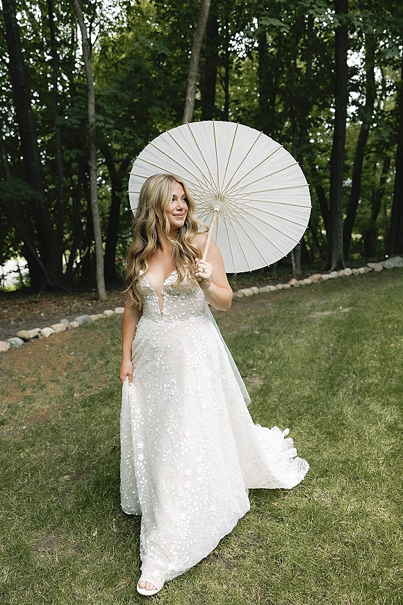 Bridal portrait of a bride with parasol in a lace deep V wedding dress and veil, walking on a stone path through a wooded park