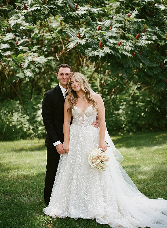 Couple portrait of bride and groom holding hands, groom hugging her as she holds a bouquet, in natural light with green trees behind