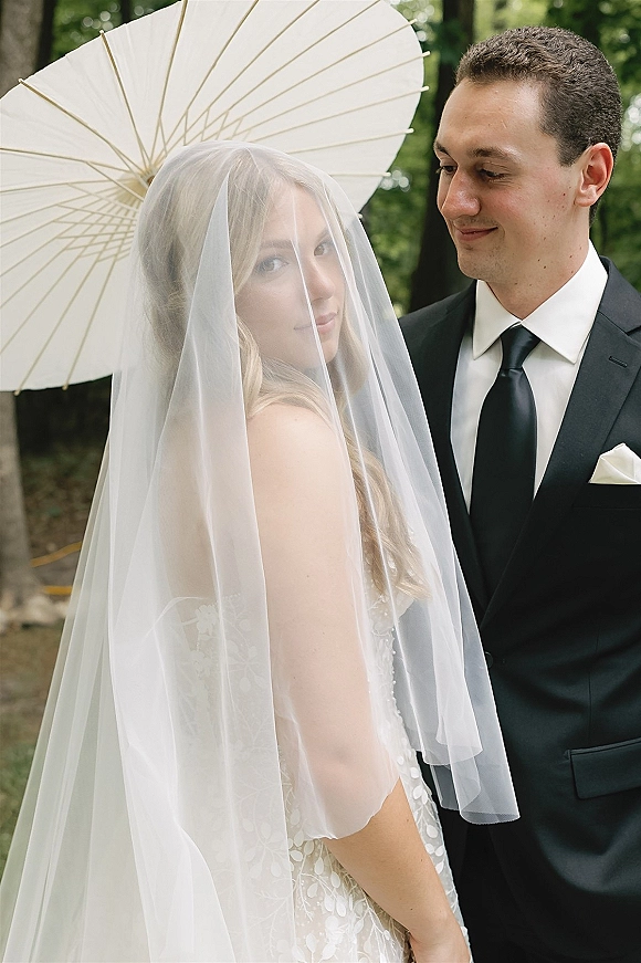 Couple portrait of bride and groom portrait with a bridal veil over her face and a white paper parasol in a lush garden backdrop