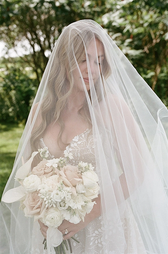 Bridal portrait of a bride with veil over her face, holding a blush and white bouquet, showing her ring in a garden setting