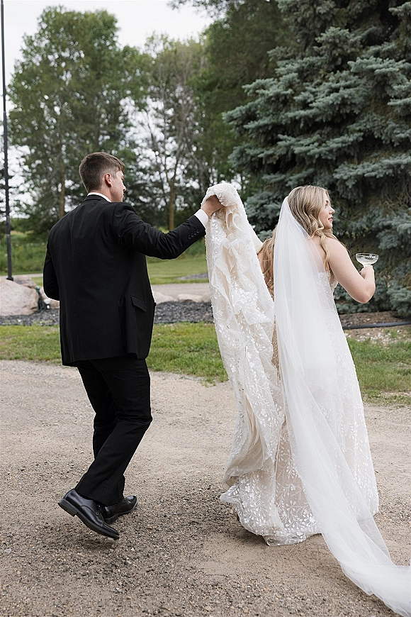Wedding couple portrait of bride and groom walking away, her lace dress and long veil flowing, holding champagne coupe on a tree-lined path