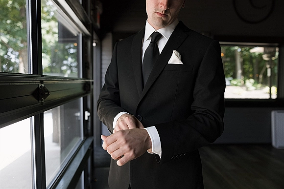 Groom portrait in window light, adjusting cufflinks on a black suit with black tie and white pocket square in a dark hallway