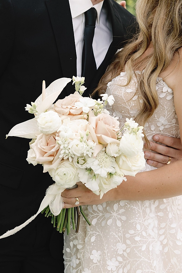 Couple portrait of bride and groom embrace, her blush rose bouquet and lace sleeves in soft natural light with greenery behind
