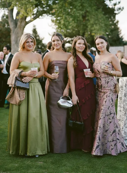 Wedding guest group in satin strapless dresses and a floral print, holding clutch purses and drinks on an outdoor lawn at cocktail hour