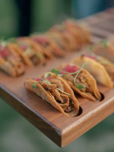 Wedding appetizers arranged as mini tacos with microgreens and diced garnish on a wooden tray against a green outdoor backdrop
