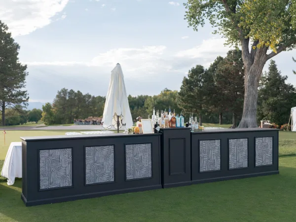 Outdoor wedding bar with a black counter, liquor bottles, glassware, garnish, and framed menu under an umbrella on a golf course lawn