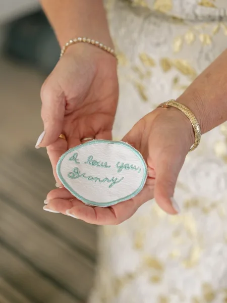 Embroidered wedding patch stitched in cursive held in bride’s hands with French manicure, wedding ring, pearl bracelet, lace dress indoors