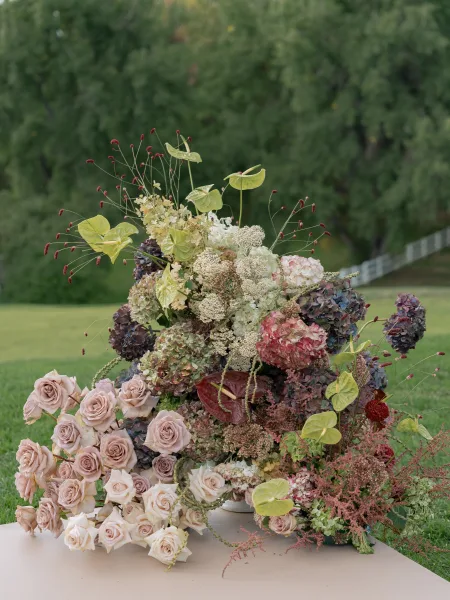 Wedding floral arrangement garden wedding centerpiece of blush roses, hydrangeas, and anthurium in a white compote vase on a lawn with trees and fence