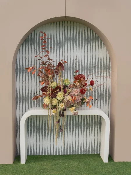 Wedding floral arrangement with ceremony florals of roses, anthurium, and hanging amaranthus on a white console table by an arched niche