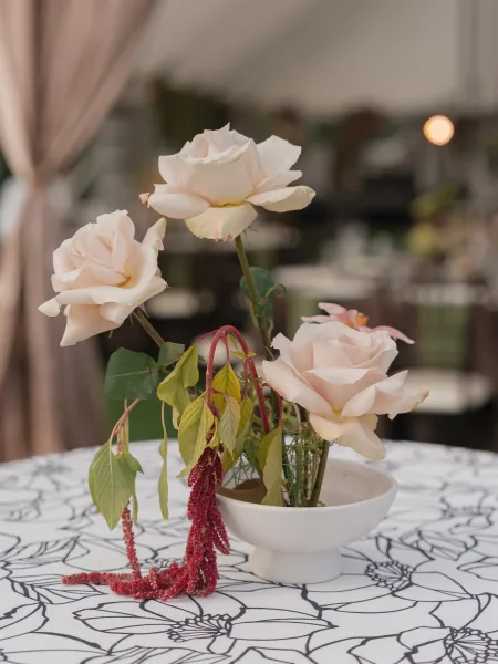 Wedding centerpiece of pale blush roses and greenery with red hanging amaranthus in a white ceramic pedestal bowl on patterned linen