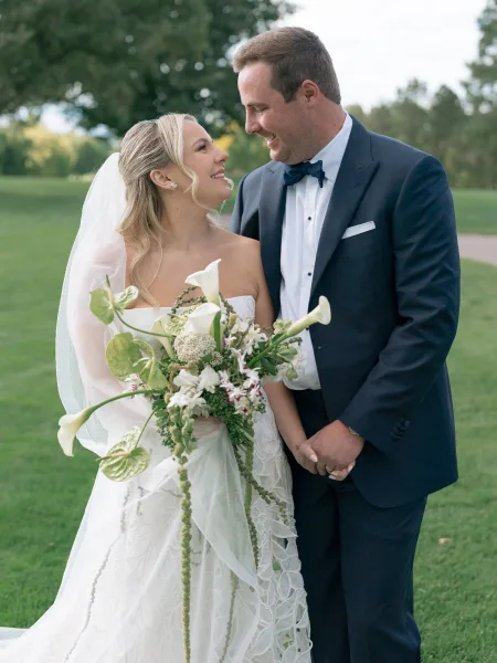 Couple portrait of bride and groom holding hands, bride with calla lily bouquet and veil, groom in navy tuxedo on a park lawn path