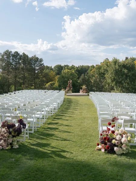 Outdoor ceremony setup with wedding ceremony seating, white folding chair rows flanking a grass aisle toward a floral platform under blue sky