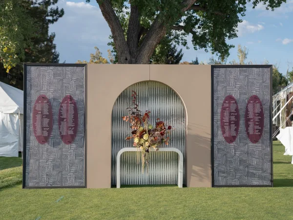 Wedding seating chart display with printed panels on a neutral arched backdrop, floral arrangement on a pedestal, set on a lawn by white tents