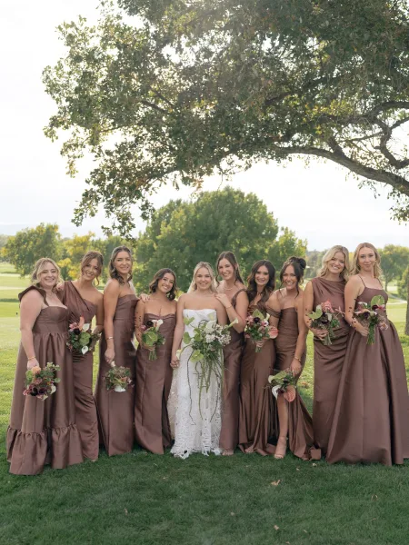 Bridesmaid group portrait with bride and bridesmaids in brown satin dresses holding calla lily bouquets under a tree canopy on a golf course lawn