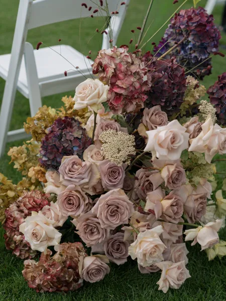 Ceremony aisle flowers with hydrangeas and roses arranged beside a white folding chair, with dried grass accents on a lawn