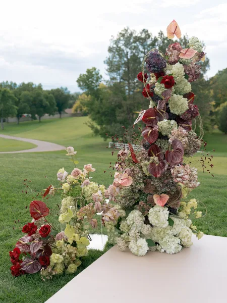 Ceremony floral arrangement of hydrangeas, roses and anthurium with greenery on a white platform set on a grassy lawn with trees