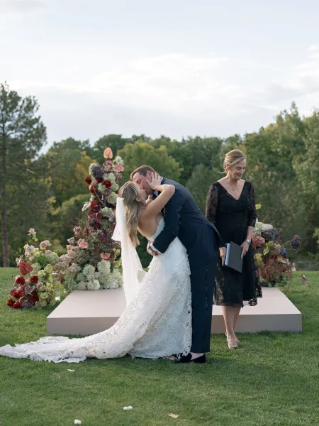 Wedding kiss as groom dips bride in long train and veil on ceremony platform, framed by floral arrangements on a grassy lawn