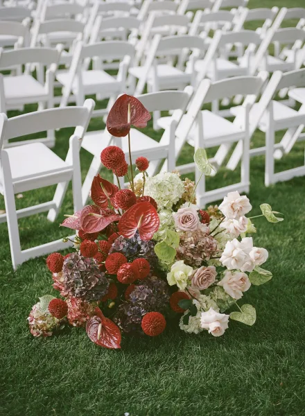 Ceremony aisle flowers in a lush wedding aisle floral arrangement with red anthurium, blush roses, and greenery beside white chairs on lawn