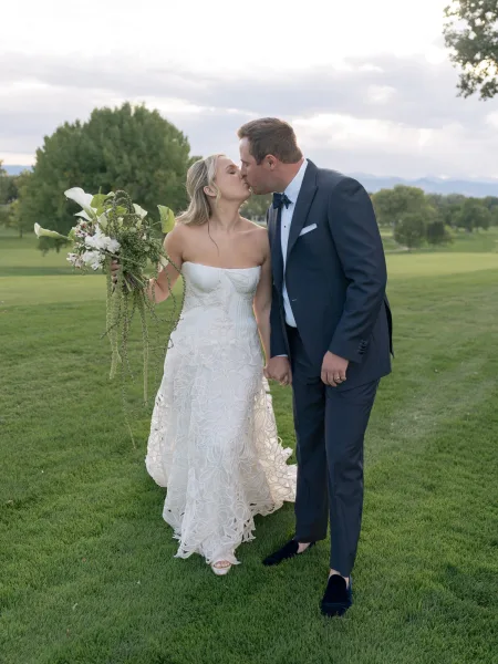 Wedding kiss portrait of bride and groom kissing, bride holding a cascading bouquet as they stand on a grassy lawn with trees and hills under clouds