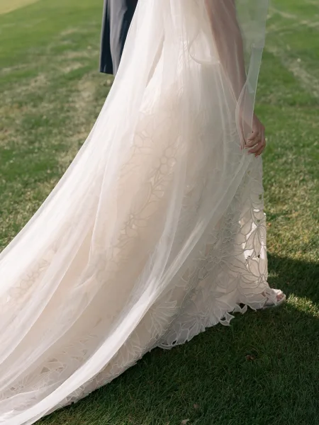 Wedding dress detail with lace wedding dress floral applique and sheer tulle overskirt, train flowing on sunlit grass lawn with strappy heels
