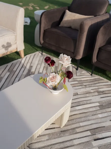 Wedding lounge seating with upholstered chairs and a white coffee table holding a small floral centerpiece on a striped rug on grass lawn