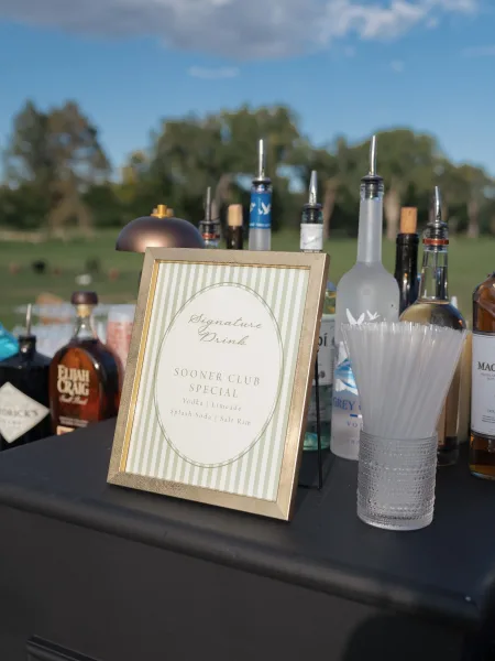 Signature drink sign in a gold frame on a bar table with cocktail bottles, pour spouts and stirrers in an outdoor field setting
