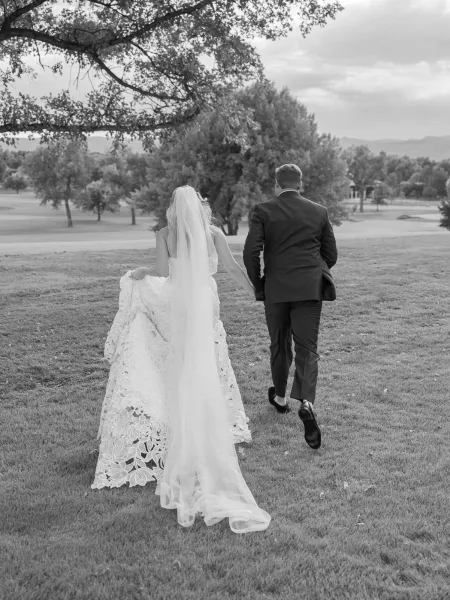 Couple walking away, bride and groom walking hand in hand as her lace train trails across a grassy lawn with mountains under cloudy sky
