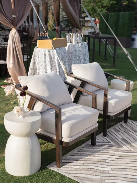 Outdoor lounge seating with white cushion chairs and pillows around a striped rug, side tables, and drink station on a grass lawn under tent poles