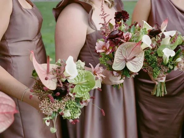 Bridesmaid bouquets with anthurium and calla lilies, dahlias and greenery held over a grass lawn beside mauve satin dresses