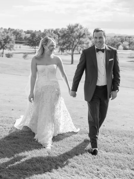 Couple portrait of bride and groom walking hand in hand on a grassy lawn with rolling hills, her veil flowing beside his tuxedo