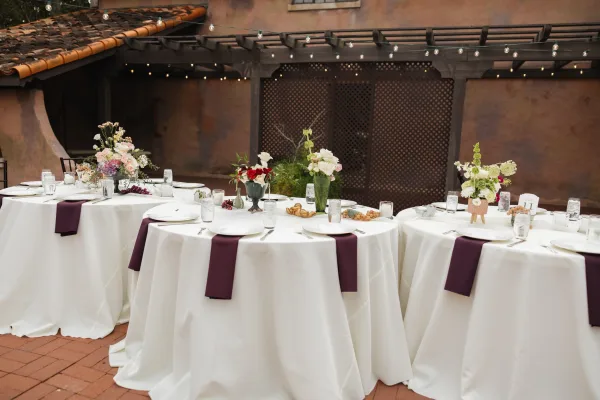 Reception tablescape with round wedding tables dressed in white cloths and plum runners, floral centerpieces and candles under courtyard string lights