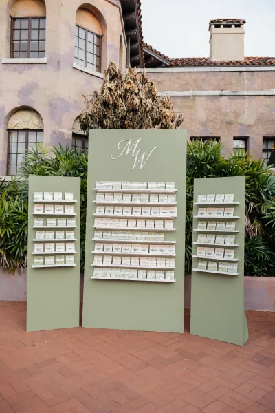 Escort card display with wedding seating chart display boards on shelves and monogram sign, set in a brick courtyard by a stucco venue with arched windows
