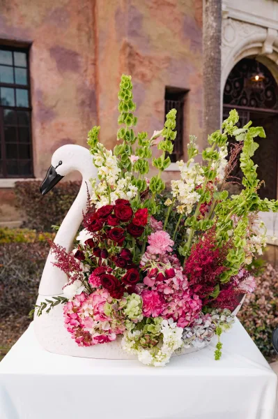 Wedding floral arrangement with swan centerpiece flowers, lush roses and hydrangea on a white tablecloth outside by a stucco wall and archway