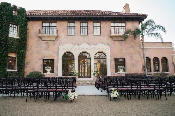 Ceremony setup with outdoor ceremony seating, a white aisle runner flanked by black chairs and floral markers in a villa courtyard lawn
