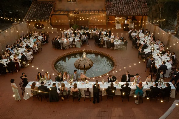Wedding reception setup with string lights over round and banquet tables, white linens, floral centerpieces and candles in a courtyard by a fountain