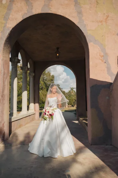 Bridal portrait of a bride in a strapless ball gown holding a rose bouquet under a columned archway with lanterns and trees behind