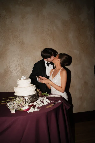 Cake cutting moment as bride in beaded gown and groom in black tuxedo kiss beside a tiered wedding cake on a styled table, textured wall behind