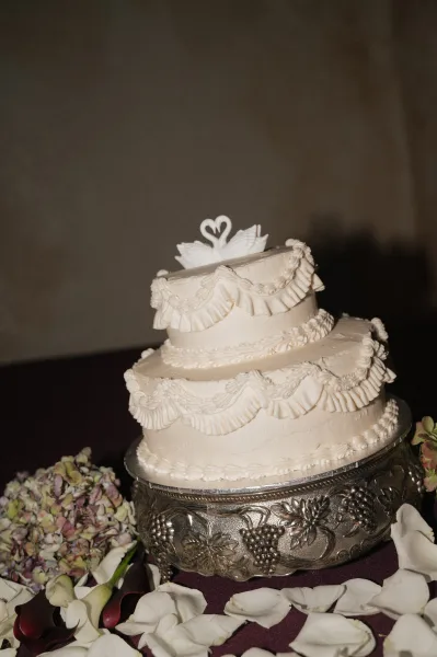 Wedding cake with white frosting ruffle piping and a swan topper on a silver stand, framed by hydrangea petals against a dark backdrop