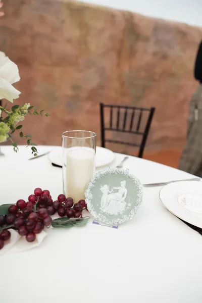 Wedding tablescape with white wedding table setting, cylinder candle centerpiece, cameo plate, red grapes, and greenery against stone wall