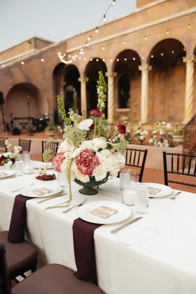 Reception tablescape with tall wedding centerpiece of white roses and pink hydrangea in a compote, burgundy runner, candles, string lights in an arched courtyard