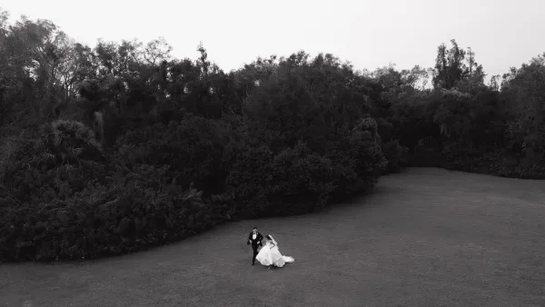 Couple portrait in black and white wedding portrait style, bride in veil and dress train holding bouquet as groom in tuxedo holds hands on a lawn