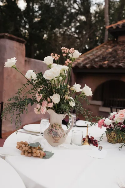Reception tablescape with wedding table centerpiece of white and pink roses in a ceramic pitcher vase, greenery runner, grapes, and brass bell outdoors