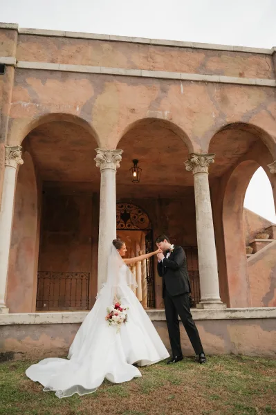 Couple portrait of groom kissing the bride’s hand as she holds a red and white bridal bouquet on steps by arched stucco columns